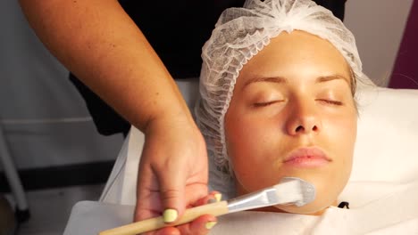 Stable-handheld-frame-right-close-up-headshot-of-a-woman-having-a-facial-done-by-a-professional-esthetician-whose-right-hand-holds-a-brush-with-the-skincare-product