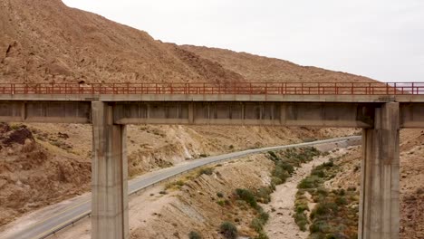 aerial-slow-backward-drone-shot-under-empty-train-bridge,-revealing-the-mountains,-an-empty-side-road,-dry-stream-and-the-desert-in-daylight