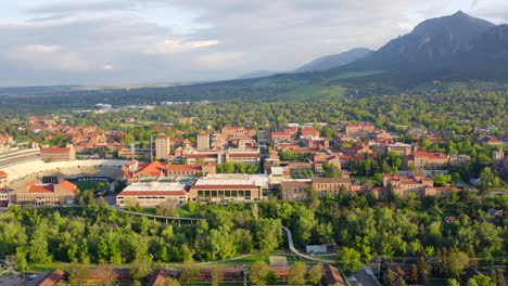 Aerial-pan-up-reveal-of-beautiful-flatiron-mountain-vista,-bright-green-trees,-and-CU-Boulder-campus-in-Boulder-Colorado-during-an-evening-sunset-with-warm-light-on-the-summer-landscape