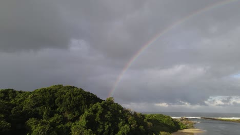 Rising-above-a-lush-green-coastal-headland-next-to-s-secluded-river-mouth-with-a-large-colorful-rainbow-over-the-ocean