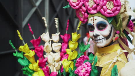 A-woman-wearing-a-traditional-outfit-and-sugar-skull-makeup-holds-colorful-flowers-at-the-Day-of-The-Dead-Parade-in-Mexico-City,-Mexico