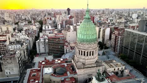 Breathtaking-aerial-shot-capturing-the-cupola-of-Palace-of-the-Argentine-National-Congress-and-Heroic-Quadriga-statue-against-downtown-cityscape-and-beautiful-orange-sun-setting-in-the-background