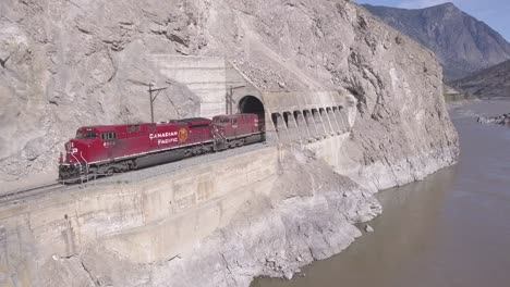 CP-freight-train-of-open-hopper-cars-emerges-from-riverside-tunnel