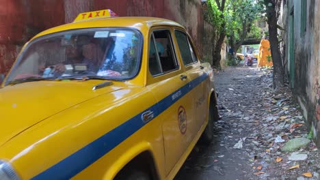 Taxi-Amarillo-Saliendo-Del-Garaje-Con-Paredes-Rojas-A-Través-De-La-Calle-De-Kolkata-En-Un-Sábado-Por-La-Mañana-Temprano-En-Un-Día-Soleado