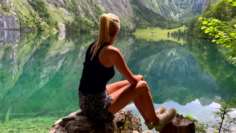 Attractive-young-woman-enjoying-crystal-clear-lake-in-nature-with-mountains-in-background---4K-Slow-motion-shot-of-Caucasian-blond-girl-sitting-on-shore-and-enjoying-wilderness