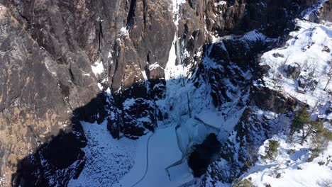 Stunning-winter-aerial-flying-over-the-cliffs,-revealing-spectacular-frozen-vøringsfossen-far-down-in-deep-canyon---Norway