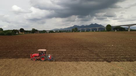 Arial-footage-of-a-tractor-ploughing-a-farmland-going-to-the-left-also-revealing-a-high-speed-railway-and-mountains-in-the-horizon-during-a-cloudy-day