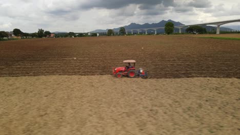 Sliding-towards-the-left-aerial-footage-of-a-hard-working-farmer-driving-red-tractor-across-farmland,-tilling-and-preparing-for-new-batch-of-plants,-high-speed-railway-network-on-horizon