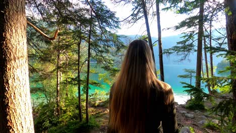 Third-person-view-of-a-blonde-girl-in-a-green-dress,-walking-through-a-forest-until-she-reaches-the-shore-of-the-famous-lake-Eibsee-in-Germany