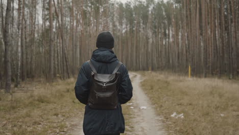Man-walking-on-forest-trail-with-a-backpack-a-jacket-and-a-cold-weather-hat,-alone-and-unrecognizable-person-seen-from-the-back
