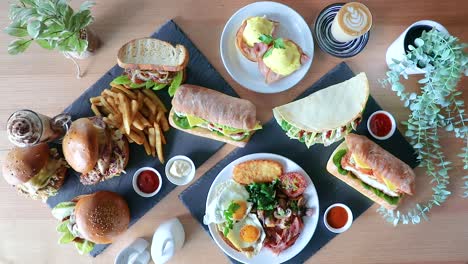 Male-hand-rotating-eggs-benedict-on-cafe-brunch-flat-lay-with-sandwiches,-burger-and-chips