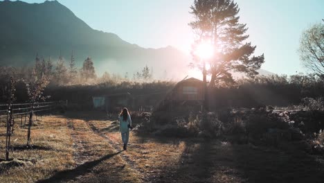 Female-photographer-with-her-camera-walking-towards-a-cabin-in-a-mountain-valley-during-a-beautiful-shiny-sunrise-day,-seen-from-the-back