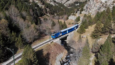 Aerial:-blue-rack-train-in-the-Pyrenees-mountains-crossing-a-bridge-and-a-river-and-going-across-a-very-narrow-valley