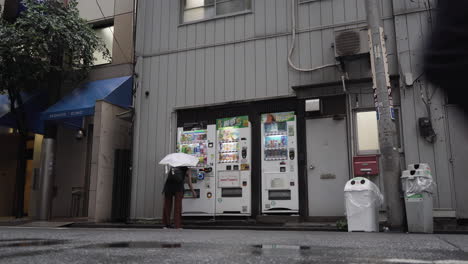 Frau-Mit-Regenschirm-Nutzt-Automaten,-Um-An-Einem-Regnerischen-Tag-In-Der-Straße-Von-Tokio-Ein-Getränk-Zu-Kaufen