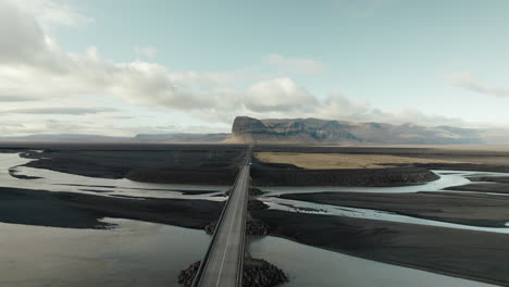 Flyover-above-bridge-on-highway-running-through-black-sand-desert-in-Iceland-towards-rock-formation