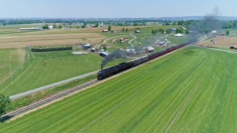 An-Aerial-View-of-a-Steam-Engine-Puffing-Smoke-and-Steam-with-Passenger-Coaches-Approaching-Passing-a-Corn-Maze-Park-and-Farmland-Countryside-on-a-Beautiful-Cloudless-Spring-Day