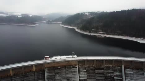 Aerial-shot-of-a-lake-created-by-the-arch-dam-Vir-in-czech-republic-with-dense-pine-forest-covered-by-snow-on-all-sides-on-a-cloudy-wintry-evening