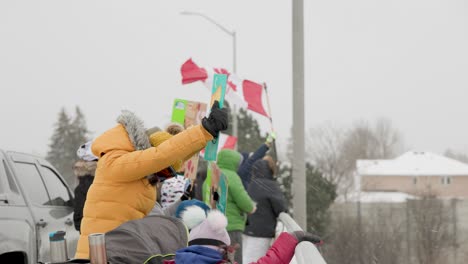 Kaukasische-Mutter-Mit-Kindern-Hält-Ein-Schild-Inmitten-Einer-Menge-Demonstranten,-Die-Pappschilder-Halten-Und-Kanadische-Flaggen-Schwenken,-Während-Starker-Wind-Und-Schnee-über-Die-Autobahnbrücke-Wehen