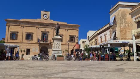 Low-angle-and-slow-motion-of-man-in-bicycle-at-Piazza-Europa-square-on-Favignana-island-with-Town-Hall-in-background,-Sicily