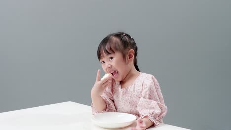 Fröhliches-Kleines-Asiatisches-Mädchen-Reibt-Sich-Die-Augen,-Lächelt-Und-Genießt-Es,-Zu-Hause-Knusprigen-Buttertoast-Zu-Essen