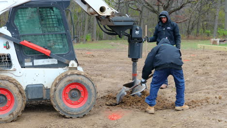 Three-construction-workers-working-with-shovels-and-hydraulic-auger-mounted-on-skid-steer-loader-to-dig-a-post-hole-for-the-frame-of-new-barn-construction