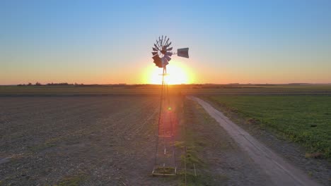 Sunset-landscape,-cinematic-aerial-tracking-shot-capturing-old-fashioned-windpump-spinning-in-the-middle-of-arable-land-in-La-Pampa,-Patagonia,-Argentina