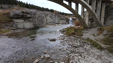 Slo-Mo-aerial-shot-approaching-Lundbreck-falls-while-flying-underneath-an-arch-bridge-in-southern-Alberta,-Canada