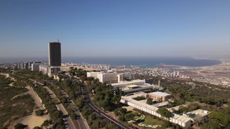 Flight-to-Haifa-city-around-a-tall-building-on-a-hill-surrounded-by-trees-and-roads,-with-the-harbor-and-the-sea-in-the-background,-Israel