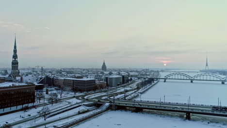 Aerial-backwards-shot-of-Riga-City-during-snowy-winter-day-in-the-morning---Frozen-Daugava-River,-driving-cars,bridge-and-cathedral-during-sunrise---Riga-Capital-of-Latvia