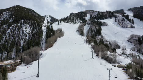 People-skiing-down-beautiful-hill-side-in-winter-resort-during-snowfall,-aerial-drone-view