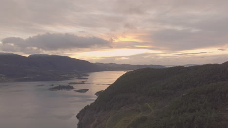 Panoramic-aerial-flight-over-forested-mountain-next-to-fjord,-fire-sunset