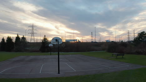 Sun-setting-behind-a-basketball-net-and-vehicles-driving-past-on-a-nearby-highway-with-large-utility-towers-in-the-background-in-slow-motion