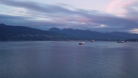 Aerial-Sunset-Medium-Wide-Shot-Flying-Over-Pacific-Ocean-Towards-North-Shore-Panning-Right-To-Reveal-Cargo-Ships-And-Downtown-City-Skyline-In-Vancouver-British-Columbia-Canada