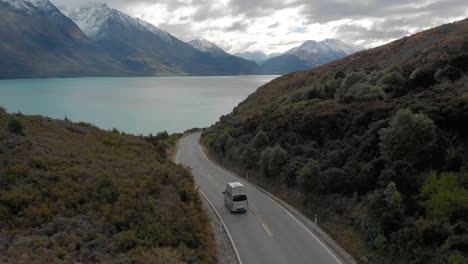 SLOWMO---Motorhome-driving-in-the-mountains-along-the-coast-of-Lake-Wakatipu,-Queenstown,-New-Zealand-with-mountains-fresh-snow-in-background---Aerial-Drone