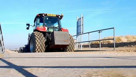 Close-up-low-angle-of-red-tractor-passing---heading-down-concrete-ramp-to-sandy-beach