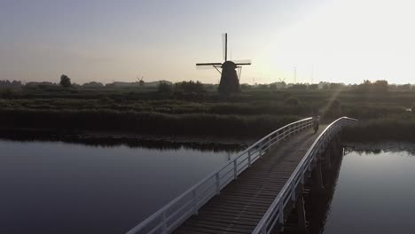Eine-Drohnenaufnahme,-Die-Nach-Rechts-Schwenkt,-Um-Ein-Modell-Herum,-Das-Auf-Einer-Brücke-Läuft-Und-Bei-Sonnenaufgang-Auf-Holländische-Windmühlen-In-Den-Niederlanden-Blickt