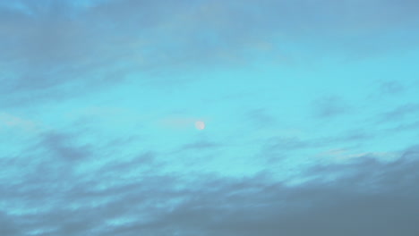Zooming-out-on-a-small-moon-in-the-sky-surrounded-by-clouds,-taken-early-evening