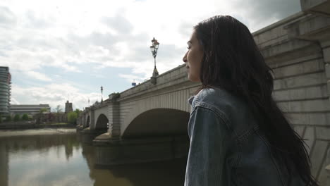 Hispanic-Latina-tourist-looking-at-the-river-Thames-and-Putney-Bridge-in-London,-smiling-and-having-a-great-time