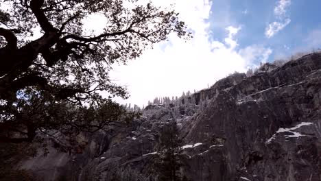 Langsam-Nach-Oben-Rotierende-Aufnahme-Der-Von-Bäumen-Gesäumten-Klippen-Des-Yosemite-Parks-Vor-Blauem-Himmel-Und-Flauschigen-Weißen-Wolken