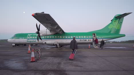 Close-wide-shot-of-people-boarding-an-Aer-Lingus-airplane-in-Cork,-Ireland-at-dawn-with-moon-in-the-background