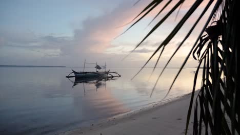 Traditionelles-Philippinisches-Bangka-Boot-Auf-Ruhigem-Wasser-In-Der-Abenddämmerung,-Vom-Strand-Hinter-Einer-Palme-Aus-Gesehen