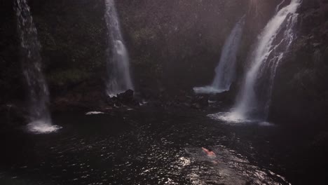 Drone-Shot-of-a-man-swimming-towards-four-gushing-waterfalls-with-mist-spraying-all-around