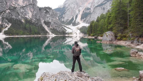 Backwards-revealing-drone-shot-of-a-male-model-standing-on-a-rock-overlooking-lake-Braies-in-the-Dolomites,-Italy