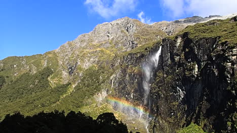 Stetige-Weitwinkelaufnahme-Des-Wasserfalls-Des-Rob-Roy-Gletschers-In-Neuseeland,-Der-Im-Wind-Fällt-Und-An-Einem-Sonnigen-Tag-Mit-Blauem-Himmel-Einen-Regenbogen-Erzeugt