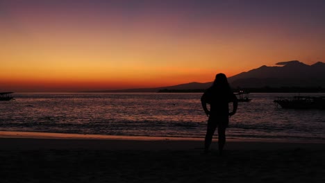 Silhouette-of-girl-standing-on-exotic-beach-in-front-of-dark-sea-surface-reflecting-a-low-light-of-twilight-orange-sky-over-Indonesian-horizon