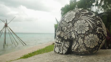 Mexican-sculpted-skull-from-side-with-wooden-tipi-structure-on-lagoon-in-background