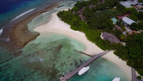 Eine-Insel-Auf-Den-Malediven-–-üppige-Insel-Auf-Weißem-Sand-Mit-Gästehäusern,-Holzsteg-Und-Am-Ufer-Angedockten-Booten,-Umgeben-Von-Aquablauem-Meerwasser-–-Luftaufnahme