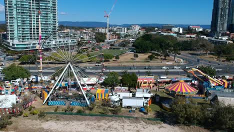Aerial-view-of-a-beach-side-carnival-next-to-a-busy-main-road-with-buildings-in-the-background