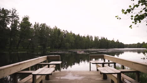 Push-in-of-lake-by-forest-and-wooden-bridge-with-tables-on-cloudy-day