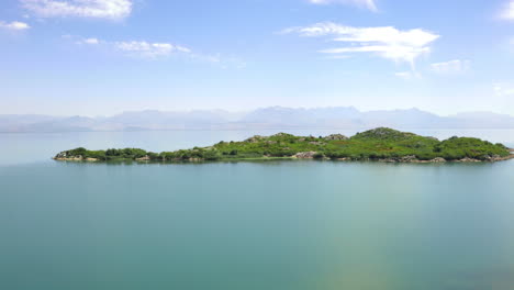 A-small-island-in-lake-Skadar-in-Montenegro-covered-with-lush-vegetation-and-rocky-outcrops,-washed-by-clear-blue-waters-on-a-sunny-day,-hazy-mountains-on-the-horizon,-aerial-zooming-view-4K